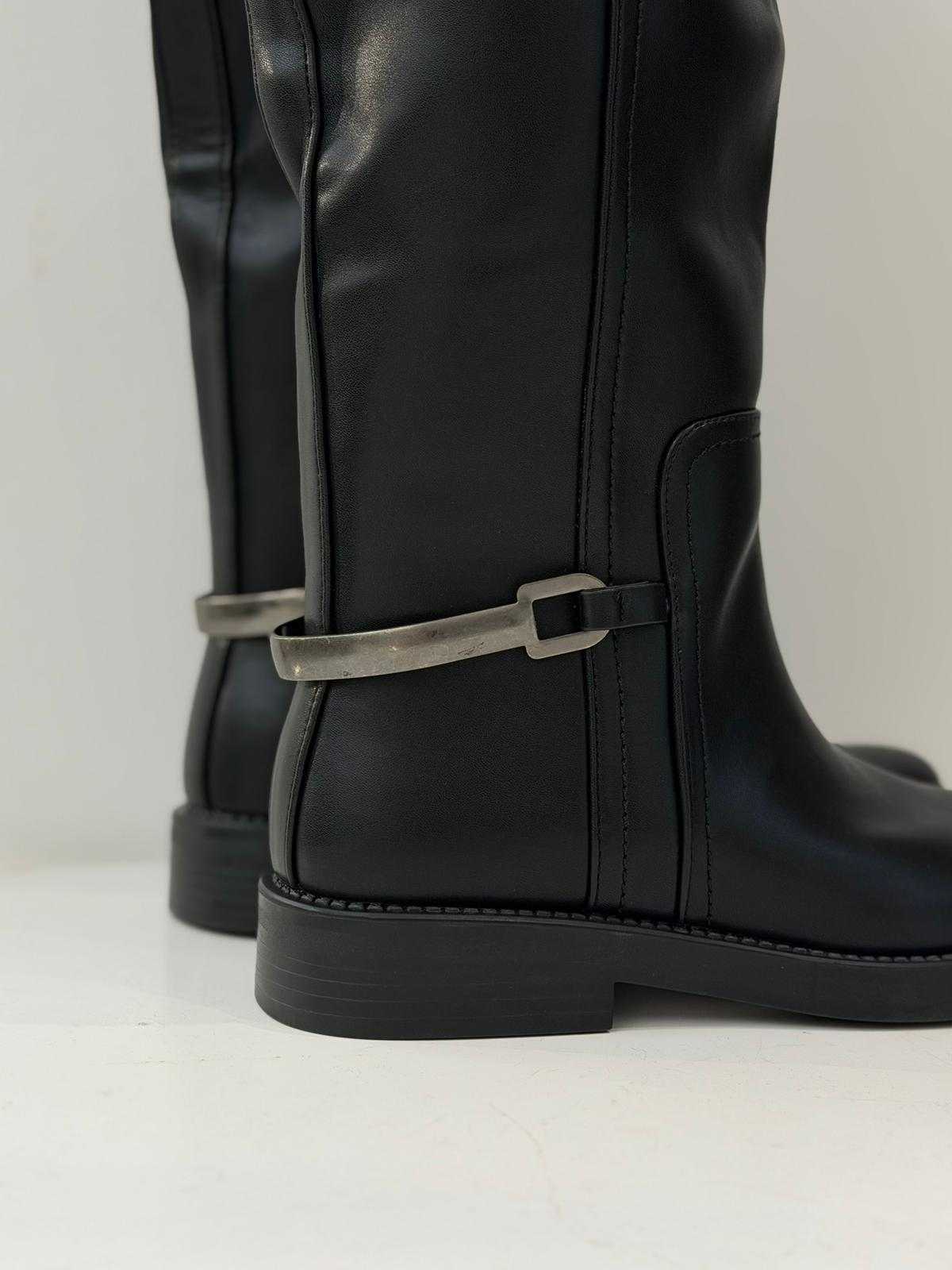 Women’s black knee-high flat boots with metal strap detail and smooth finish
